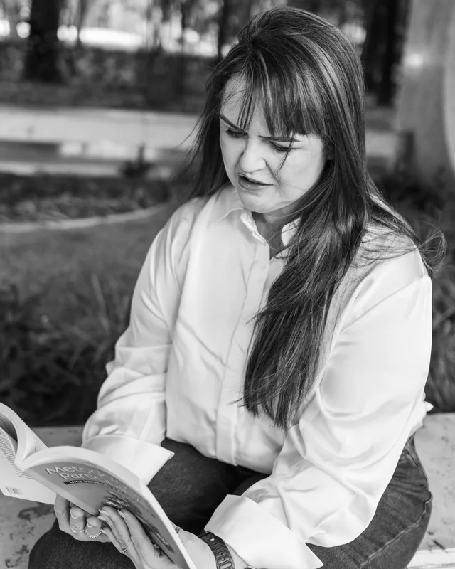 Ana Carla Moura, em preto e branco, sentada ao ar livre lendo um livro